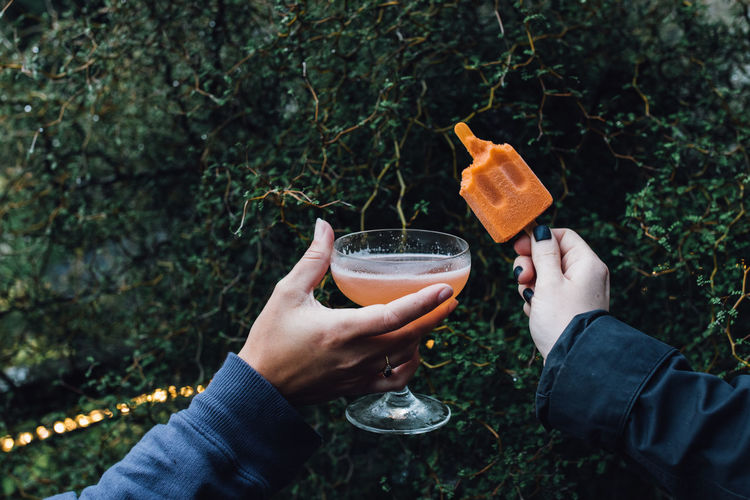 Two hands holding up orange cocktail and popsicle, cheers outdoors