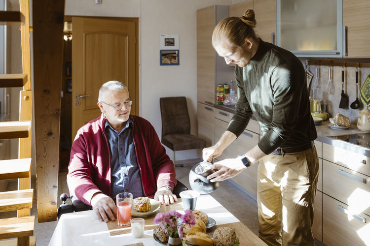 Young male caretaker pouring coffee for retired senior man with disability during breakfast at home