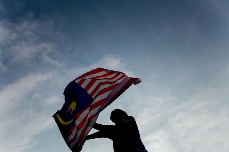 Low angle view of man waving malaysian flag against sky
