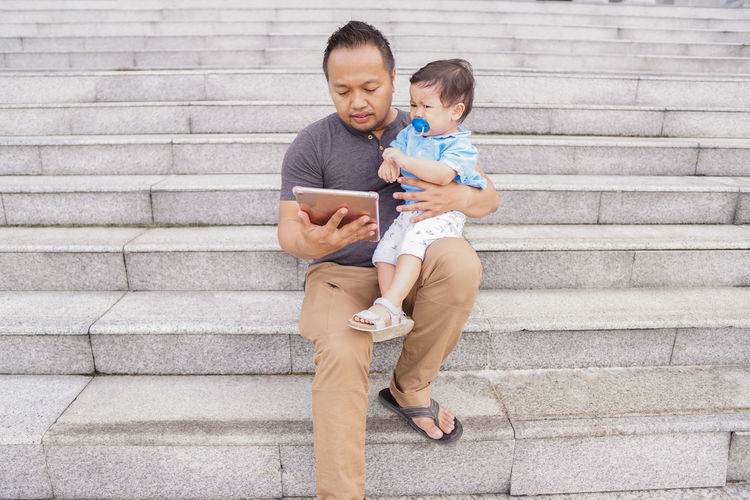 Man using digital tablet while holding baby sitting on staircase outdoors