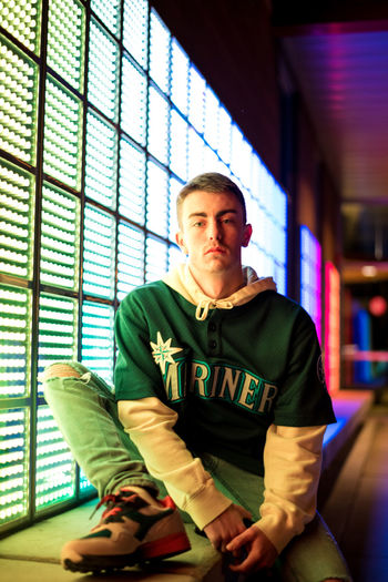 Portrait of serious young man sitting by illuminated building at night