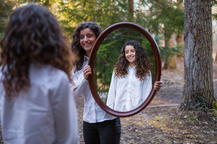 Smiling woman holding mirror in front of sister at forest