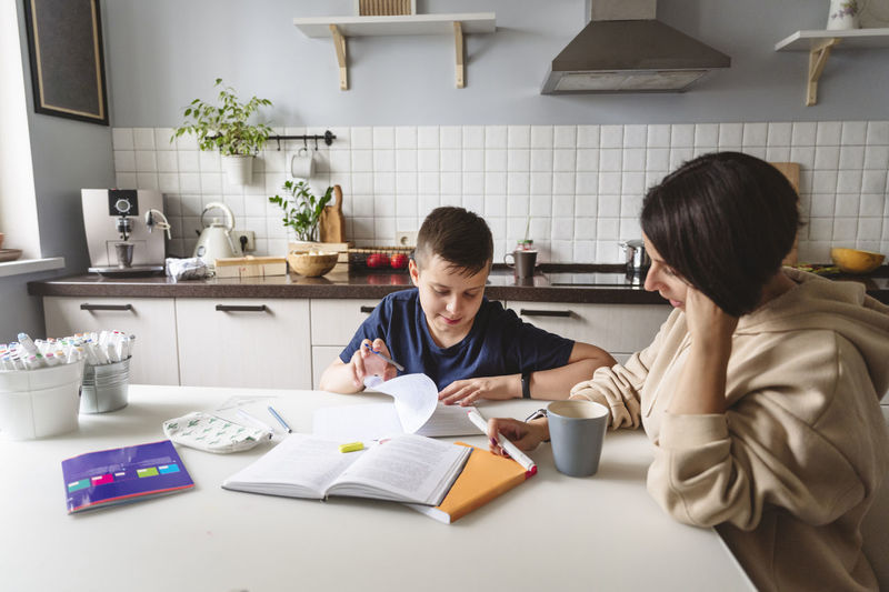 Mother assisting son in homework while sitting at dining table in kitchen at home