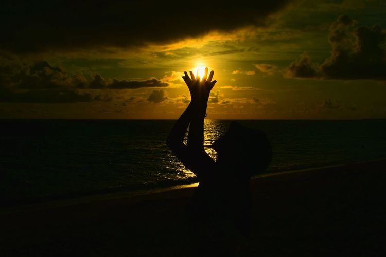 Optical illusion of silhouette woman holding sun at beach against sky during sunset