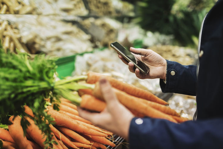 Midsection of woman using phone while buying carrots in supermarket