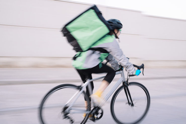 Side view of anonymous female courier with thermal bag riding bike on street road while delivering food in city, motion blur