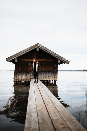 Unrecognizable female traveler in warm clothes standing on wooden plank pier near shabby shed located at lake in gloomy autumn day