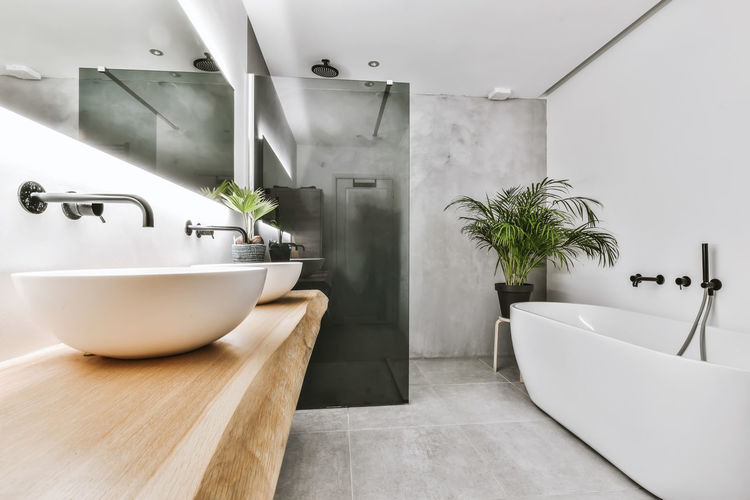Oval shaped bathtub and shower cabin in modern spacious illuminated bathroom with white ceramic sinks and wooden cabinet