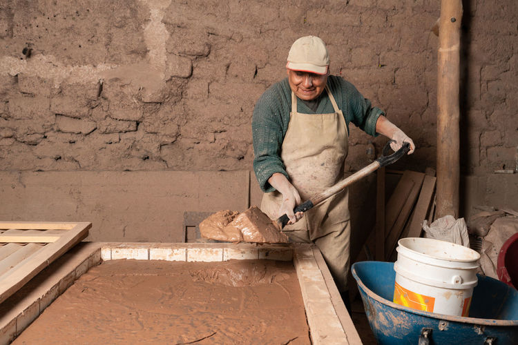 Focused peruvian male worker with dirty hands in uniform carrying shovel with plaster near plastic bucket while working in ceramics workshop