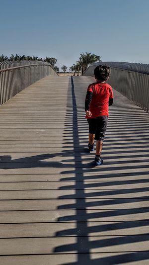 Rear view of boy walking on footbridge against sky
