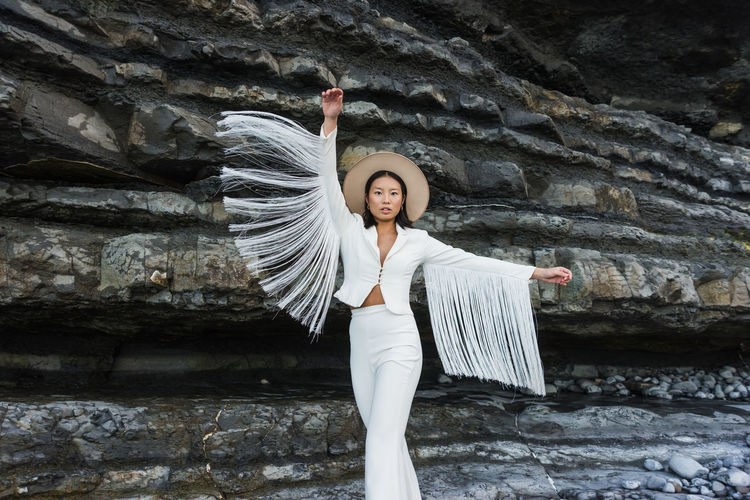 Elegant young slim female model in white suit with wings shaped tassel sleeves and hat outstretching arms while standing against rough rocky formation