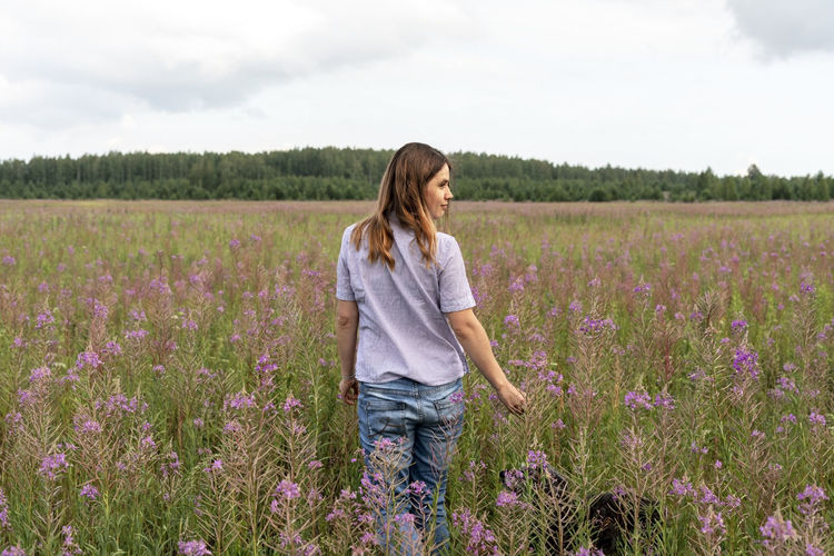 Rear view of young blond woman walking on field among pink flowers of fireweed with fluffy dog