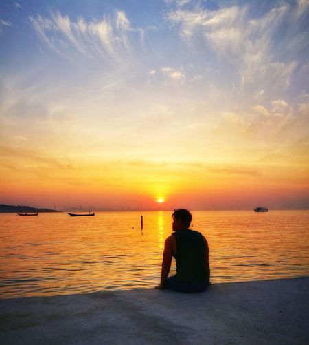 Man sitting in sea against sunset sky