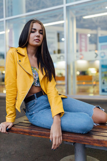 Young long haired female in trendy yellow leather jacket and ripped jeans looking at camera while sitting on bench against glass window of modern building