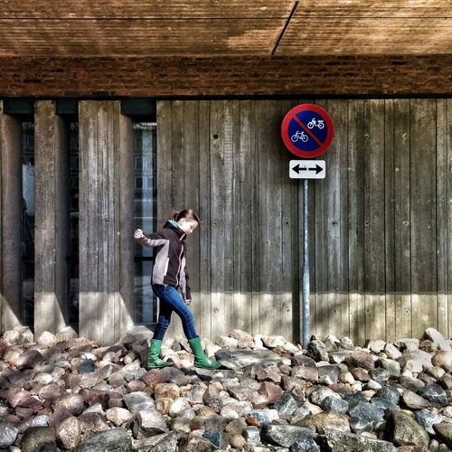 Full length of girl walking on stones by road sign against wooden wall