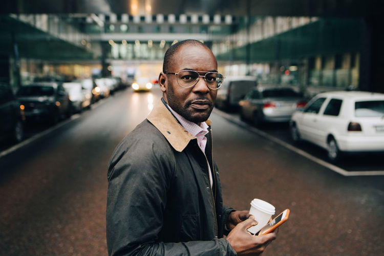 Portrait of businessman with coffee using mobile phone while standing on street in city