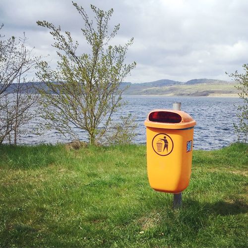 Garbage can on grassy field against lake