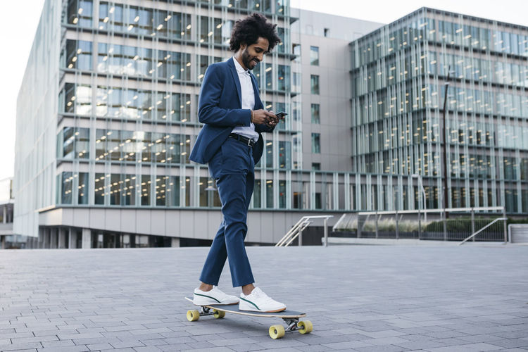 Spain, barcelona, young businessman riding skateboard and using cell phone in the city