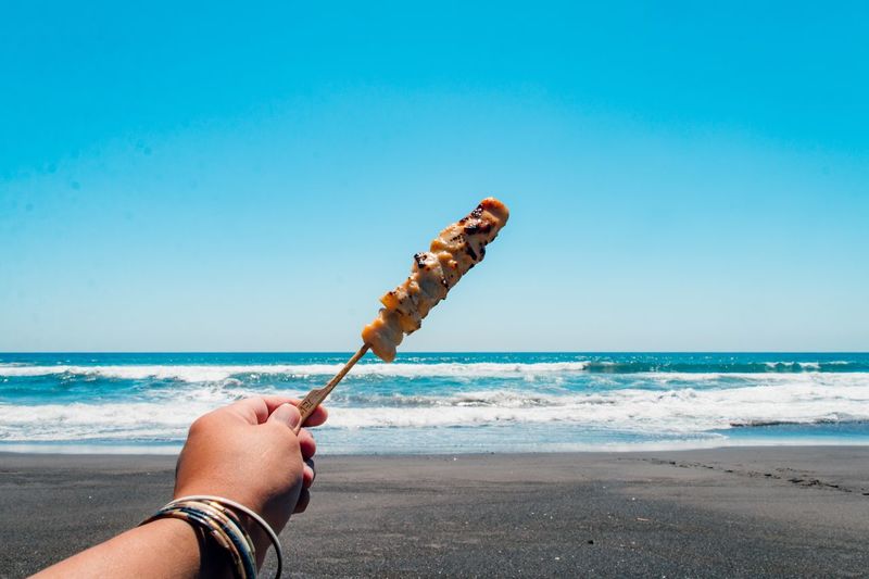 Man holding yakitori on beach against clear sky and sea