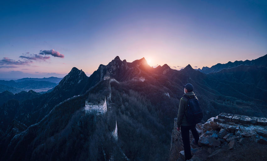 Man with backpack looking at view while standing on cliff against mountain and sky during sunset