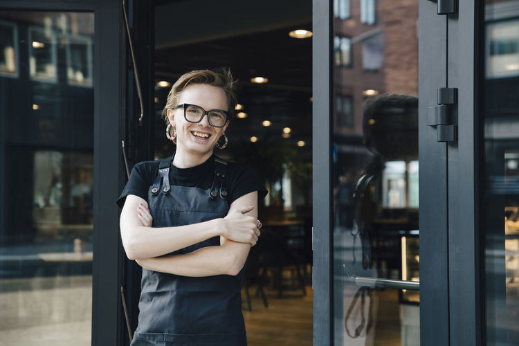 Portrait of smiling androgynous owner standing with arms crossed at coffee shop entrance