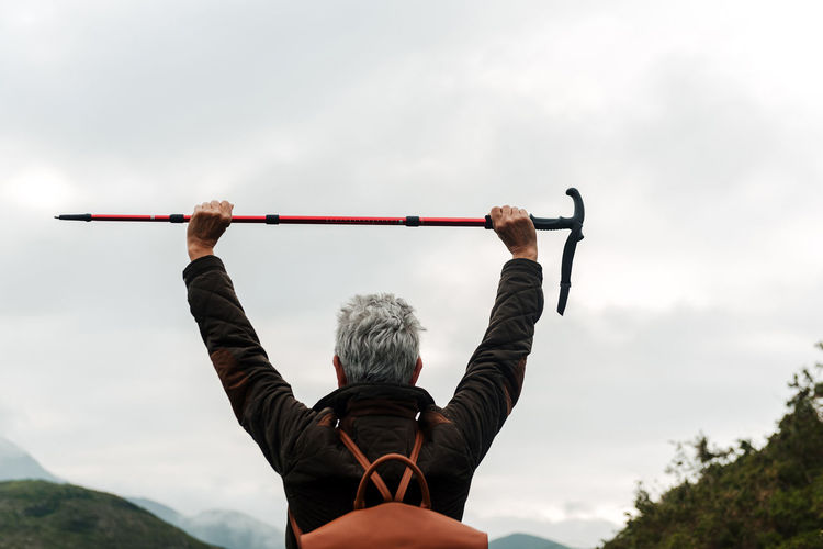 Back view of anonymous aged woman with backpack holding walking stick in raised arms against cloudy gray sky while exploring nature