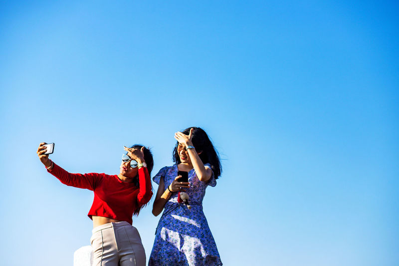 Low angle view of female friends taking selfie while standing against clear blue sky