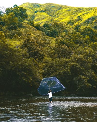 Fisherman throwing fishing net over river against green mountain