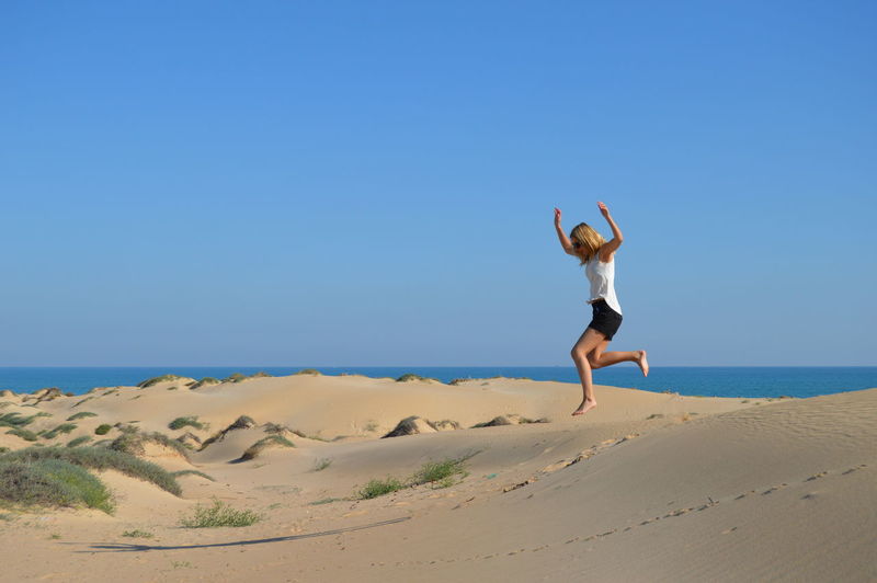 Full length of happy woman jumping on sand at beach against clear blue sky