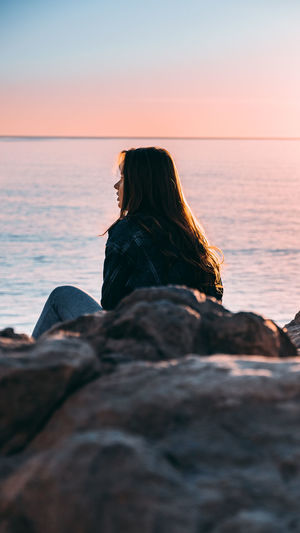 Midsection of woman sitting by sea against sky during sunset