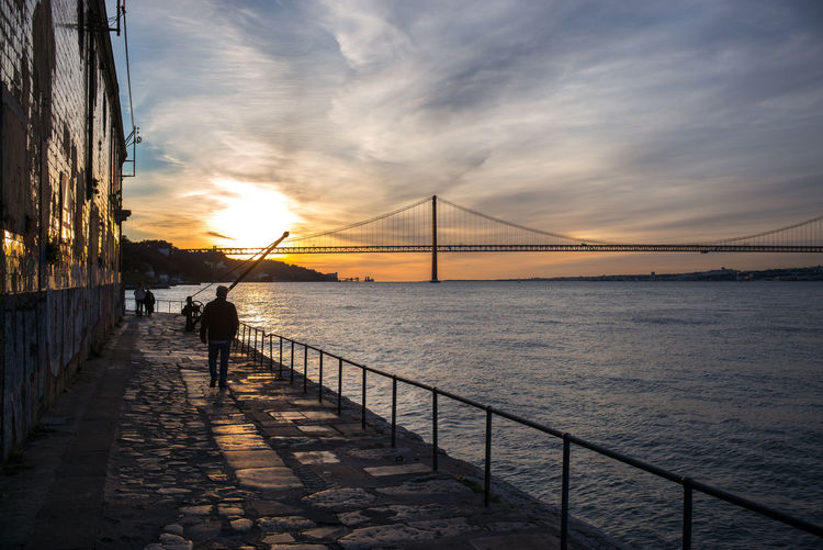 Silhouette man standing on suspension bridge over sea during sunset