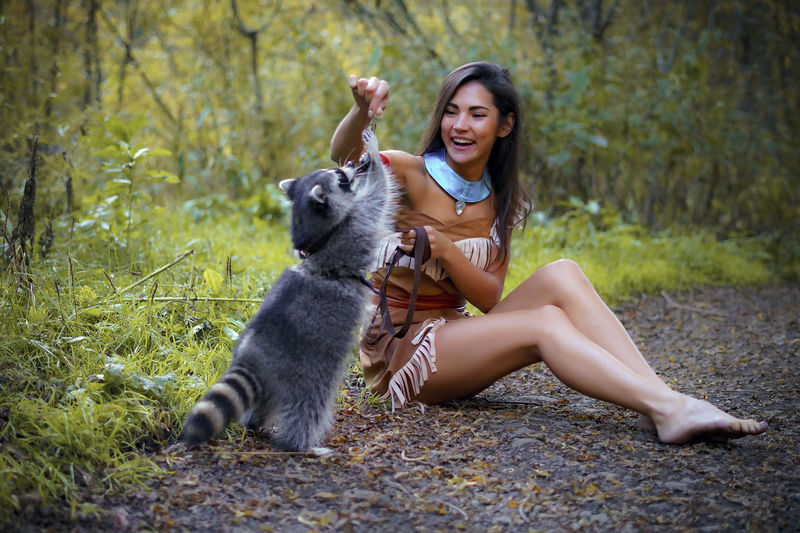 Young woman in traditional clothing playing with raccoon in forest