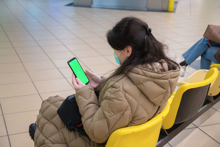A young woman in a warm down jacket with green screen smartphone sits at subway