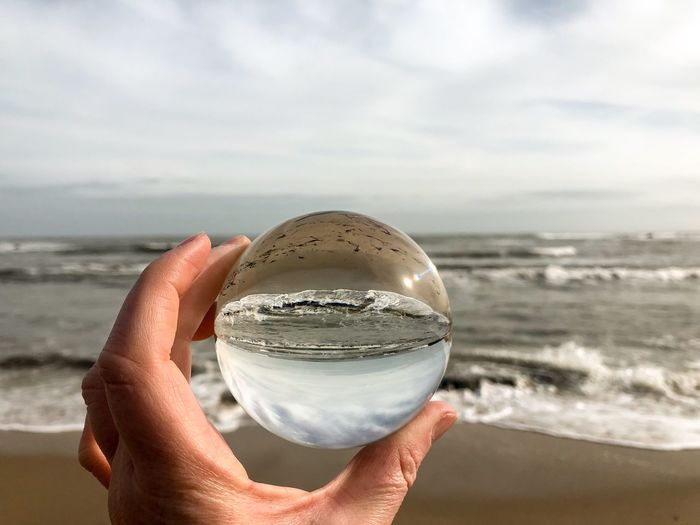 Cropped hand holding crystal ball by sea against cloudy sky