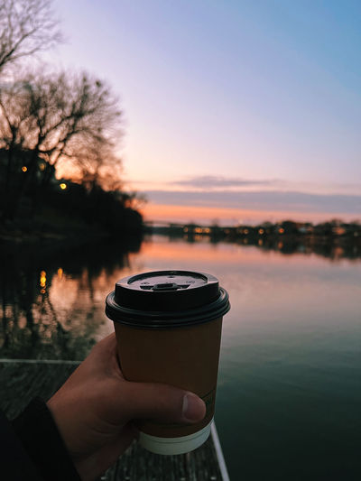 Cropped hand of person with coffee cup at sunset river