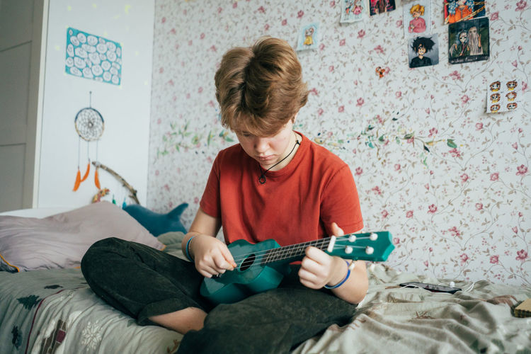 A teenage girl sitting on a bed plays the ukulele.
