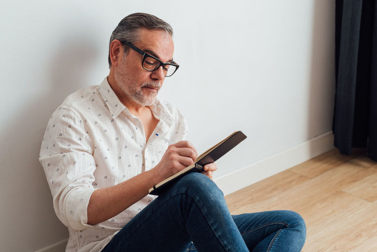 Focused mature male in casual wear and eyeglasses sitting on parquet at home and writing down thoughts in diary