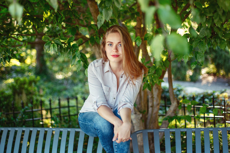 Portrait of smiling young woman sitting on bench against trees