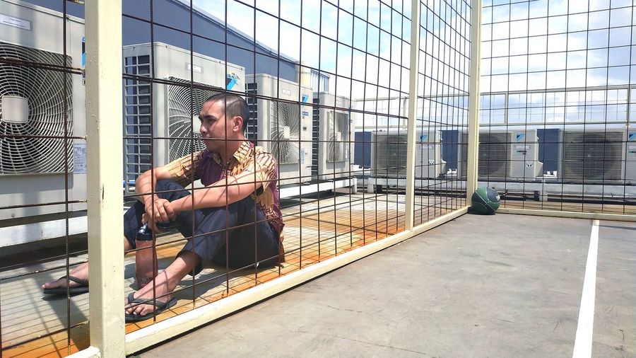 Man sitting against air conditioners seen through fence
