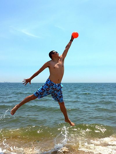 Full length of shirtless man jumping while catching ball over shore during summer