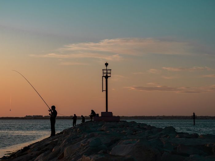 People fishing in sea against sky during sunset