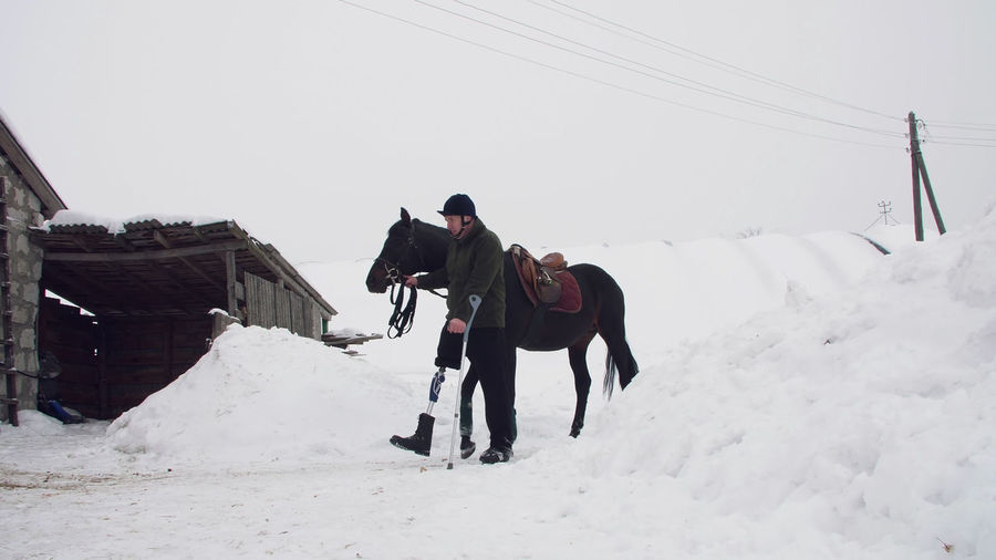 Snowy winter, disabled man jockey leads, holding with reins a black horse on the way. 