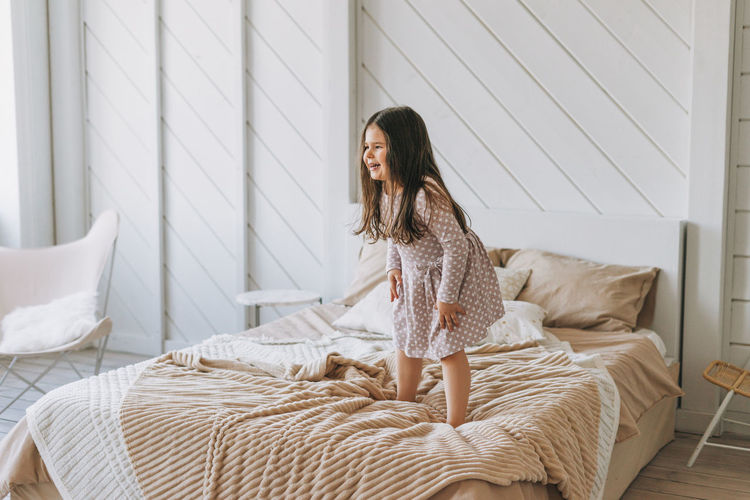 Cute long hair little girl jumping on parents bed at bright room, scandinavian interior