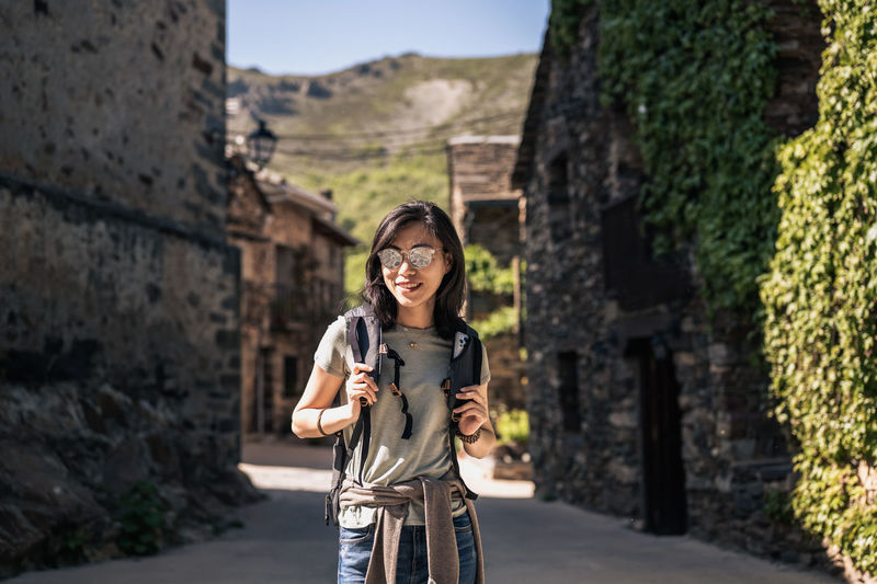 Asian female tourist with backpack standing on street with old stone buildings in province of guadalajara in spain on sunny summer day
