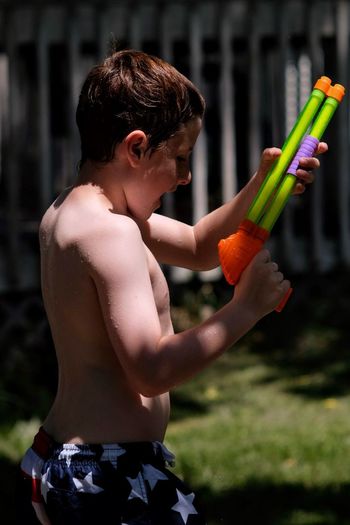 Side view of shirtless boy holding squirt gun in yard
