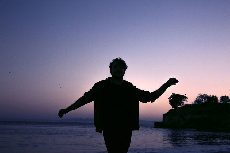 Silhouette man with arms outstretched standing by sea against sky during sunset