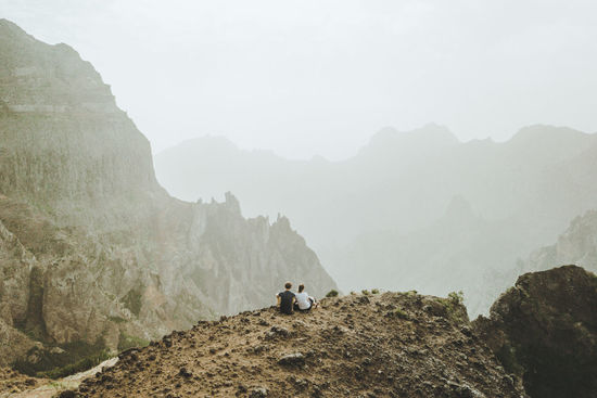 Rear view of people sitting on cliff by mountains against sky