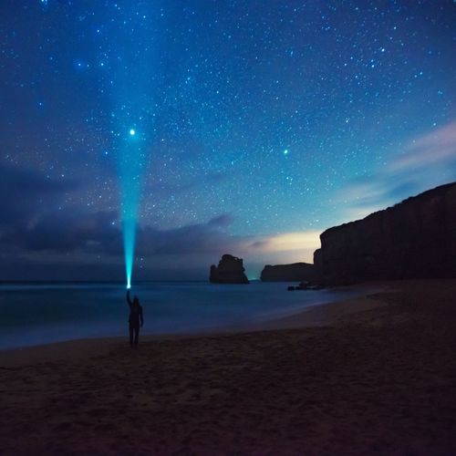 Silhouette man pointing flashlight towards star field while standing on sea shore