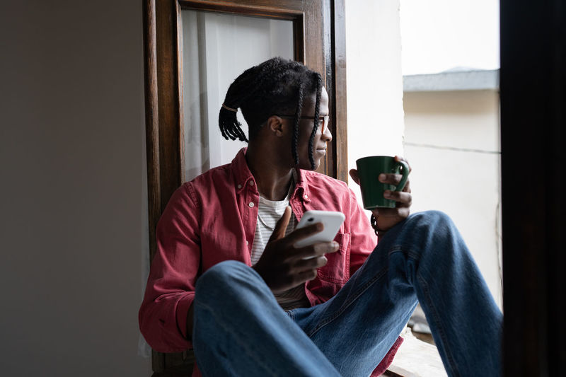 Young smiling african guy holding smartphone and cup of tea relaxing in silence by window at home