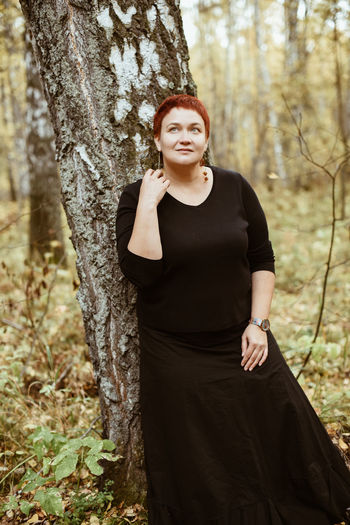Middle-aged woman of 30-40 years old in autumn forest stands by tree, pensive, dreamy.